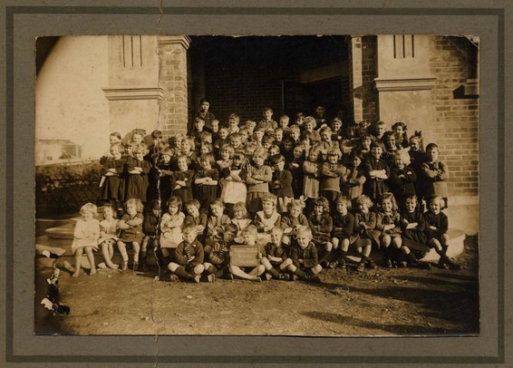 Black-and-white group photo: Foxton School primary school pupils, July 1924. Description: infants’ pupils are seated at the front with older pupils standing behind, in front of the entrance to the school building.  There are about 60 pupils, all with their arms crossed. Citation: Foxton Historical Society. Kete Horowhenua. CC BY-NC-SA 4.0 DEED. https://horowhenua.kete.net.nz/item/db0af8c7-5d95-4fb0-a5f2-7170e2648e3d