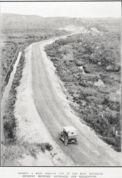 Black-and-white photo: Closing a Most Serious Gap in the Main Metalled Highway Between Auckland and Wellington. 1925. Description: Show a motor car in the foreground travelling away from the camera on a metalled road, which is raised by earthworks above the surrounding scrubland. Fencing runs along each side of the road. A road cutting can be seen in the distance as the road turns left in front of background hills. Citation: Auckland Weekly News, 30 April 1925, p. 39. Auckland Libraries Heritage Collections AWNS-19250430-39-05. https://kura.aucklandlibraries.govt.nz/digital/collection/photos/id/244136