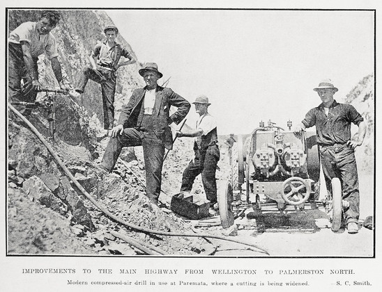 Black-and-white photo: Improvements to the Main Highway From Wellington To Palmerston North. Photographer, S. C. Smith. 1926. Extended caption: Modern compressed-air drill in use at Paremata, where a cutting is being widened. Description: Five workmen stand facing the camera in the foreground, all but one wearing hats, one man even wearing a suit jacket and waistcoat. Three men are standing on the bank of the cutting. The compressor for the drill is mounted on a low carriage that stands on the road; one man stands to the right of it, resting his hand upon it.  Citation: Auckland Weekly News, 25 March 1926, p. 50. Auckland Libraries Heritage Collections AWNS-19260325-50-03. https://kura.aucklandlibraries.govt.nz/digital/collection/photos/id/249827