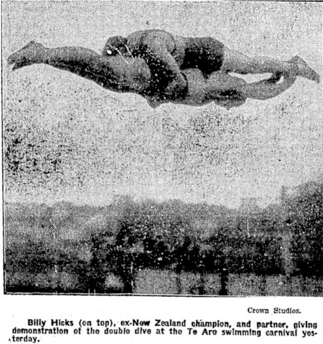 Black-and-white photo: A Double Dive. 1927. Photographer, Crown Studios. Extended caption: Billy Hicks (on top), ex-New Zealand champion, and partner, giving demonstration of the double dive at the Te Aro swimming carnival yesterday. Description: An action shot of two men in mid-dive, both divers facing downwards and head to toe. The upper diver is holding the waist of the lower diver, who is holding the ankles of the upper diver. Citation: Evening Post, 1 February 1927, p. 7. https://paperspast.natlib.govt.nz/newspapers/EP19270201.2.29.1