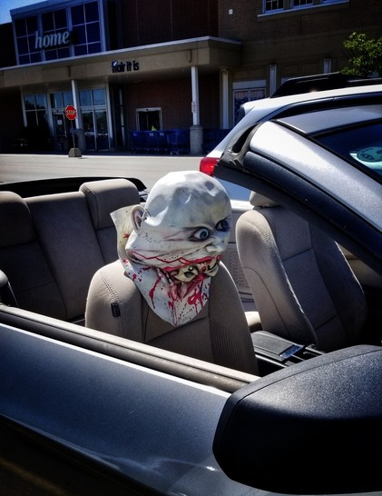 An open top convertible parked in a store lot on a sunny day. There is a scarry Halloween-ish mask over the headrest of the passenger seat.