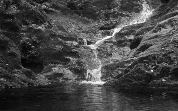 A small waterfall cascading over moss-covered rocks into a still pool. The image is low-key monochrome.