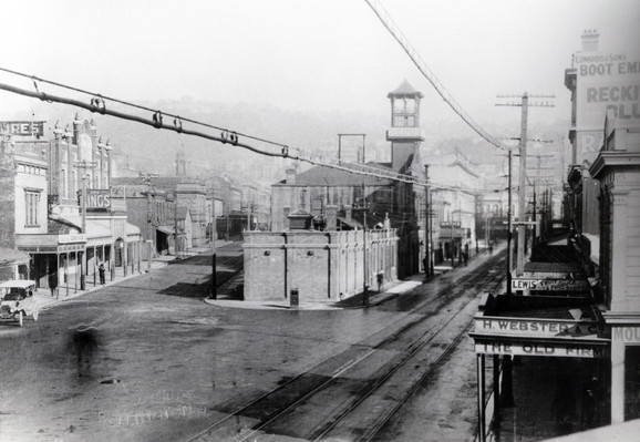 Black-and-white photo: Manners Street. ca. 1920. Photographer, Zak Studios. Description: The ‘Market Hall’ site between Manners and Dixon Streets. Taken prior to the demolition of the ‘old’ fire station (being used here as Flockton’s Variety Store). Now the site of Te Aro Park (formerly known as Pigeon Park). The view is elevated three storeys or so; we see hard-sealed streets to right and left, the right-hand street with two parallel tram tracks. There are power and/or telephone cables just overhead going in the direction of the tram tracks. Hills are indistinctly visible in the background through haze. Citation: Wellington City Libraries, Reference No. 50010-404. https://wellington.recollect.co.nz/nodes/view/8761
