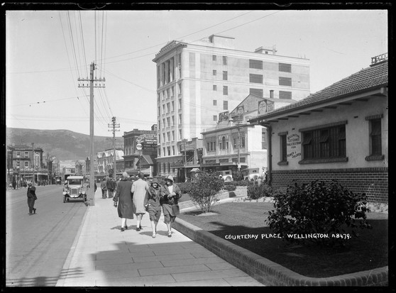 Black-and-white photo: Courtney Place, Wellington. 1920s. Photographer, Frederick George Radcliffe. Description: Looking on a sunny day southeast along Manners Street (left) towards the intersection with Taranaki Street and Dixon Street (right) and Courtenay Place (distance) showing buildings on right including: Wellington City Council Public Rest Room for Women and Children (foreground); Kings Theatre; Pram House; Hope Gibbons Ltd.; Belcher’s Buildings importers of leather goods; Fuller’s Vaudeville; on left: Hotel Argonne (on the corner of Taranaki Street) with pharmacy on ground floor; other buildings; pedestrians on footpath (BrE pavement, AmE sidewalk; centre foreground); and crossing road; cars parked (partly obscured right); truck (left foreground). The slopes of the town belt are to the left in the far background. Citation: Auckland Libraries Heritage Collections 35-R1786. https://kura.aucklandlibraries.govt.nz/digital/collection/photos/id/274855