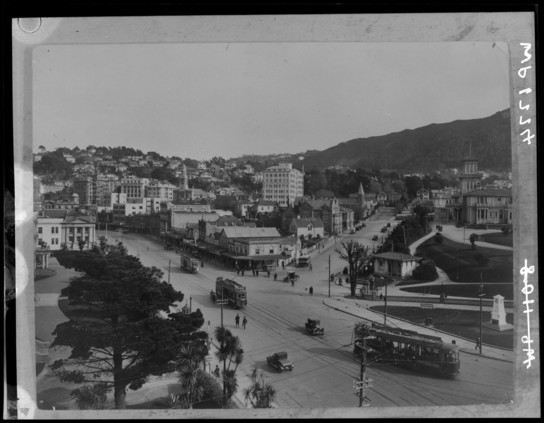 Black-and-white photo: Corner of Bowen Street and Lambton Quay. 1920s. Description: Elevated view of Bowen Street and Lambton Quay, prior to construction of the Cenotaph. Trams pass Parliament grounds and Bowen Street ends just past the intersection with The Terrace. Citation: Wellington City Council Archives, Reference No. 00138-1774. https://archivesonline.wcc.govt.nz/nodes/view/13001