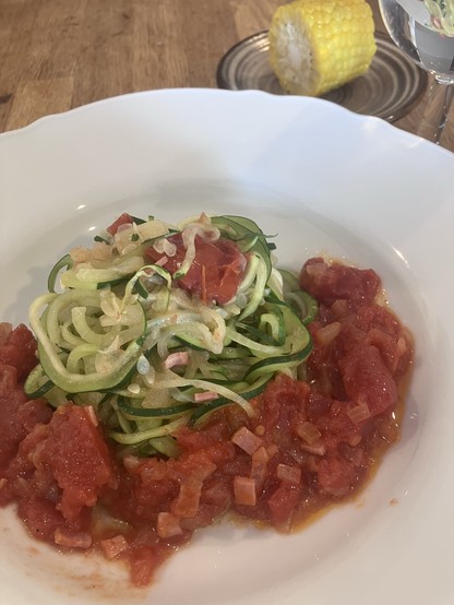 A plate of zucchini noodles topped with tomato sauce and diced vegetables. In the background, there is a half-eaten corn on the cob placed on a dish.