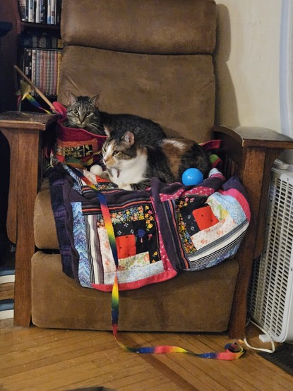 A brown tabby and a tabby calico lay on a quilt covered arm chair with wood arma.