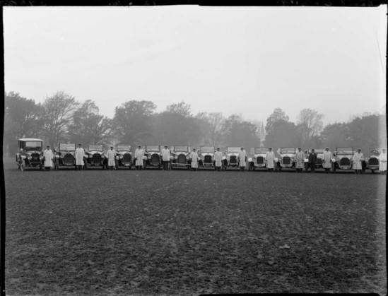 Black-and-white photo: Motor cars from David Crozier Limited, motor car importers and engineers, arranged in a field, with drivers. 1920s? Photograph taken by the Steffano Webb Photographic Studio, Christchurch. Description: Each driver is dressed in a white coat and white cap and stands to the left front of each car. Another man, possibly the business owner, is dressed in a dark suit and hat and stands in front of one of the cars towards the right of frame. Citation: Webb, Steffano, 1880-1967: Collection of negatives. Ref: 1/1-004156-F. Alexander Turnbull Library, Wellington, New Zealand. https://natlib.govt.nz/records/23192179