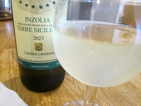 Wine glass of white wine with a bottle of Inzolia Terre Siciliano 2023 white wine beside it on wooden table