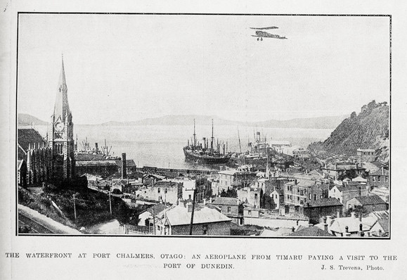 Black-and-white photo: The waterfront at Port Chalmers, Otago. 1922. Photographer, J. S. Trevena. Description: The waterfront at Port Chalmers, Otago with an aeroplane from Timaru overhead. Citation: Auckland Weekly News, 31 August 1922, p. 46. Auckland Libraries Heritage Collections AWNS-19220831-46-04. https://kura.aucklandlibraries.govt.nz/digital/collection/photos/id/239056