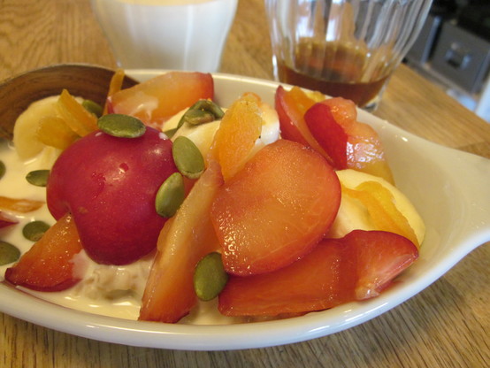 Close up shot of a dish of porrige with toppings of banana, plum, dried apricot and pumpkin seeds.  A glass of soy milk and another of tea in the background