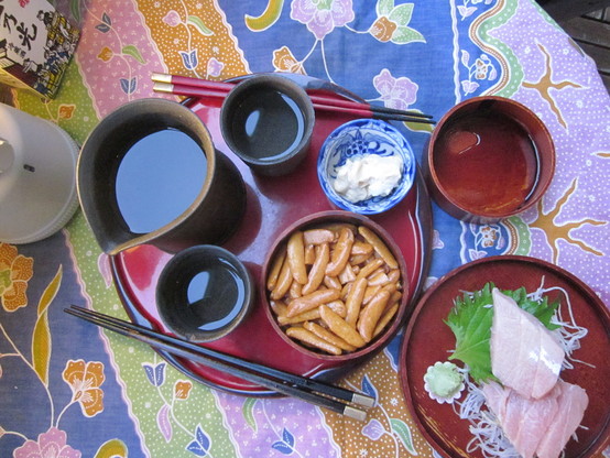 Round table on balcony with sake beaker and glasses filled with sake reflecting the evening sky, with a dish of rice crackers and a small dish of mayonnaise, and a wooden plate with sashimi and another of soy sauce for dipping, with two pairs of chopsticks