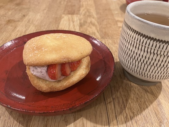 A dessert sandwich with strawberry filling on a red plate, accompanied by a cup of tea in a patterned ceramic mug.