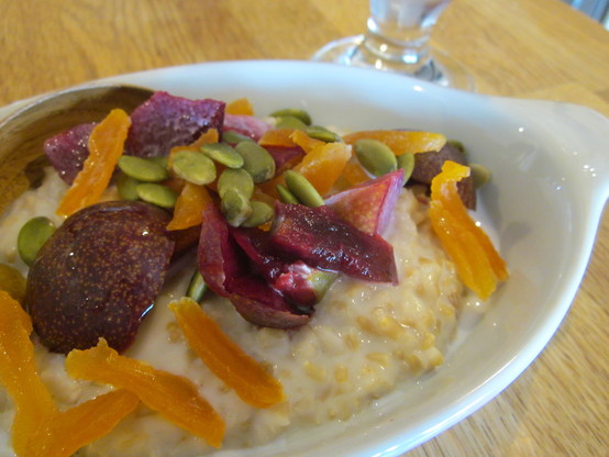 Close up on a dish of porridge topped with sliced plum, and sprinkling of dried apricot slivers and pumpkin seeds.  A glass of iced coffee in corner of frame