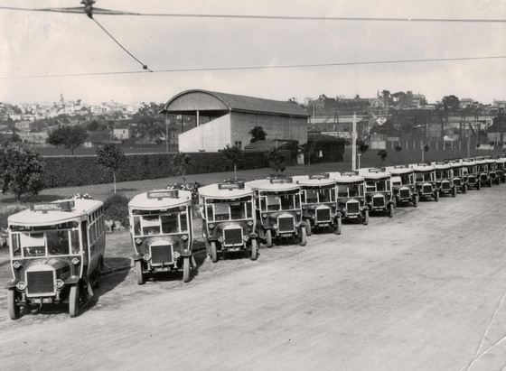 Black-and-white photo: Auckland City Corporation bus fleet. 1920s. Description: A line-up of buses on Fanshawe Street, angle-parked and facing the camera. The Victoria Park sports ground can be seen behind. Citation: Walsh Memorial Library, The Museum of Transport and Technology (MOTAT), Accession No. 08/092/249. https://collection.motat.nz/objects/95483/auckland-city-corporation-bus-fleet-1920s