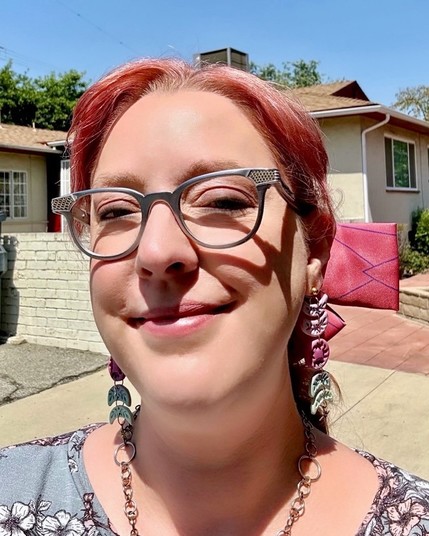 Person with pink hair and eyeglasses, wearing long decorative earrings, outside with houses in the background.