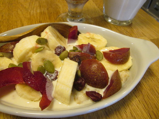 Dish of porridge with banana, plum, dried cranberry and pumpkin seed topping.  Glass of iced coffee and of cold soy milk in background