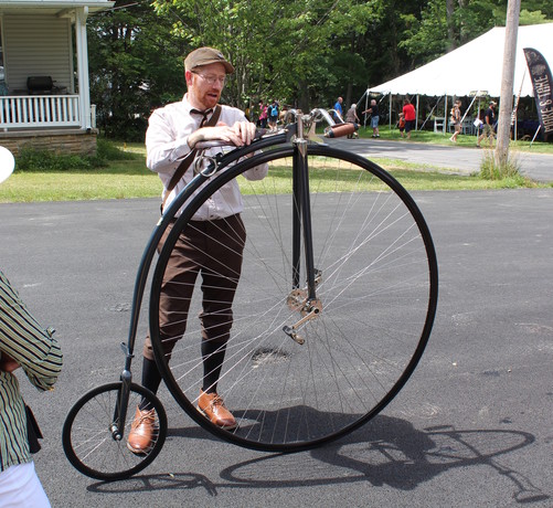 A young man in Victorian clothes stands by a high-wheeler bicycle. 