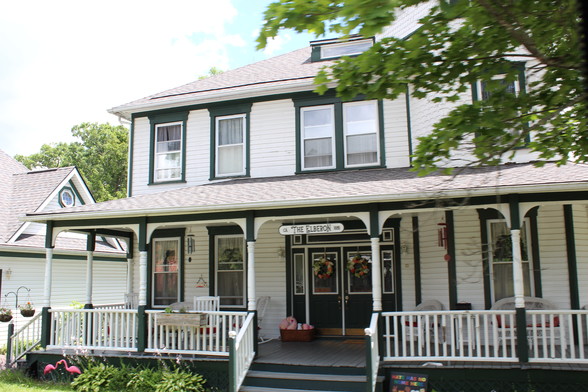A white Victorian-era house with green trim and a long, broad porch. A sign on the porch roof says, 