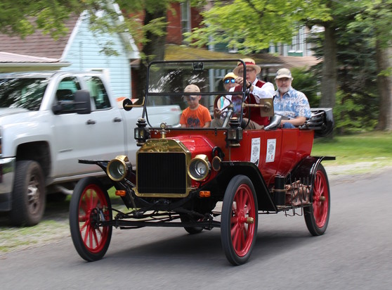 A red Model T Ford proceeds on a modern street. A man in Victorian dress, with a straw hat, drives, while the passengers are in modern dress.