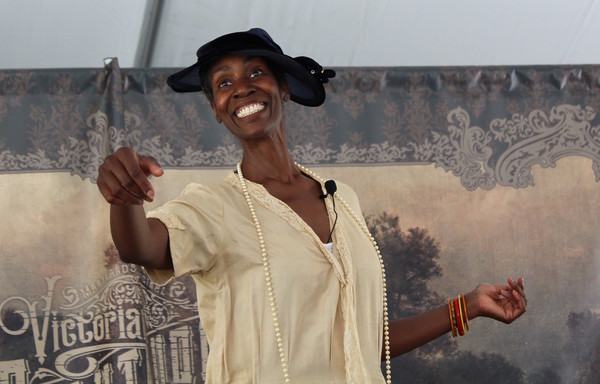 A Black woman, portraying Zora Neale Hurston, wearing a 1920s outfit, gestures with a broad smile. 