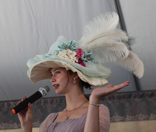 A young lady speaks into a microphone and gestures. She wears a flamboyant Victorian-style hat with roses and plumes.