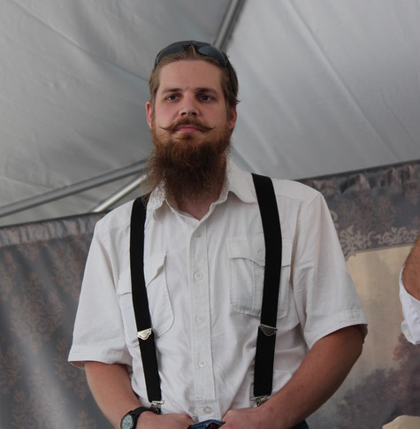 An embarrassed-looking young man, with a waxed mustache and bushy beard, in a white shirt with black suspenders, stands in a tent.