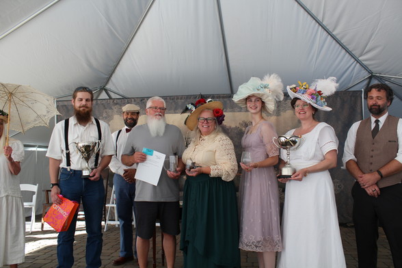 A group of people stand on a stage. Two men to the left are winners of a Best Beard & Mustache competition; three ladies are winners of the Best Victorian Hat competition. On the far right is the MC of the program. 