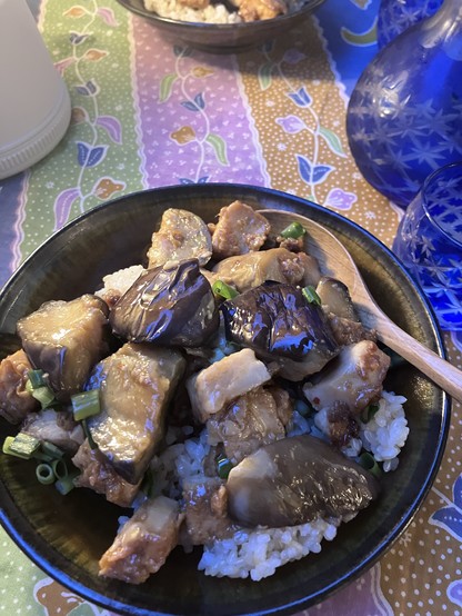 A bowl of stir-fried eggplant and tofu served over rice on a colorful tablecloth, accompanied by blue glass bottles.