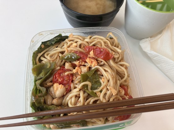Noodles with vegetables, soybeans and egg in a plastic container, served with miso soup and a beverage in white and green cups, accompanied by chopsticks and a napkin.
