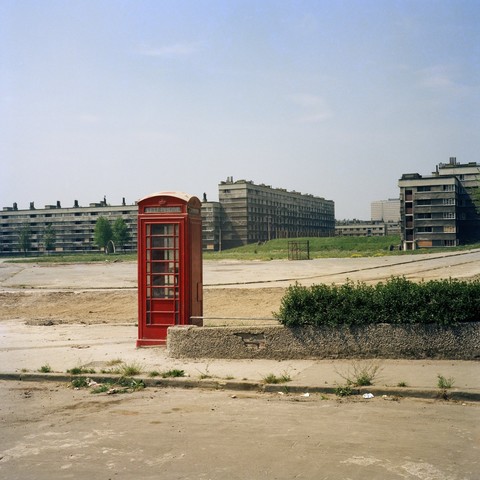 Photograph of a red telephone booth in the foreground, with a huge estate of concrete flats in the background. The booth stands next to a low wall and hedge, but otherwise appears to be isolated on an expanse of open ground, with weeds pushing through cracks in the pavement.