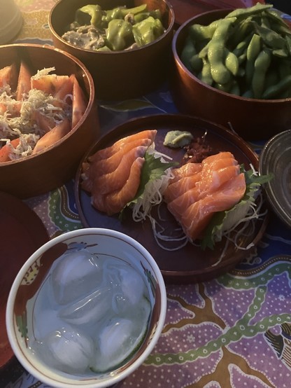 An assortment of Japanese dishes including salmon sashimi, edamame, and other vegetables, accompanied by a cold beverage with ice cubes.