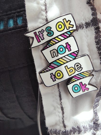 「It's OK not to be OK」という言葉が、はためいたようなリボンに書かれているデザインのピンバッジの写真。