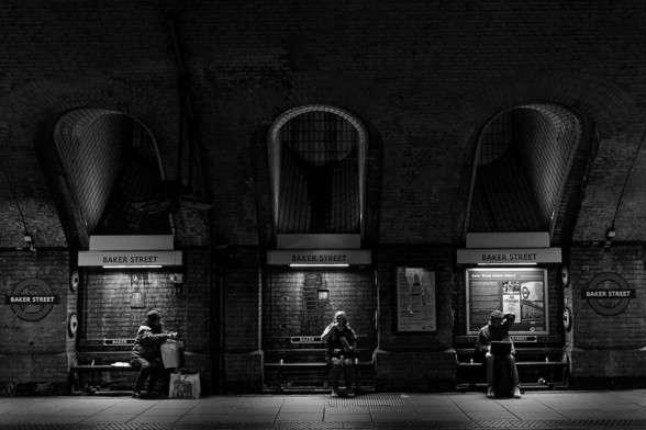 Black and white photo of the platform at Baker Street.  There are three arches inset into the wall with a bench in each.  There is one person sitting on each of the benches.