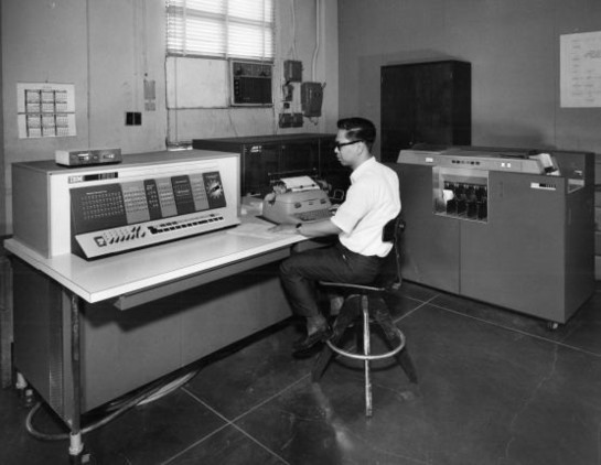 Another picture of an IBM 1620 computer with a high-speed reader-punch in the right background. An operator sits at the console typewriter.