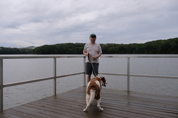 A man with a green cap standing on a deck over a lake, holding his white and brown dog with his butt to the front. A lake view and woods in the far back.