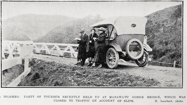 Black-and-white photo: A Dilemma: Party of Tourists Recently Held Up at Manawatu Gorge Bridge, Which Was Closed to Traffic on Account of Slips 1923. Photographer, B. Lambert. Description: Shows a man and two women, all wearing long coats and hats, standing beside a stationary car on an unsealed road. One woman is facing the camera, the others are seen in left profile. The car, facing away from the camera, is stopped just in front of a small bridge to the left. Citation: Supplement to the Auckland Weekly News, 28 June 1923, p. 44. Auckland Libraries Heritage Collections AWNS-19230628-44-04. https://kura.aucklandlibraries.govt.nz/digital/collection/photos/id/236948