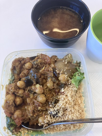 A lunch container with chickpea stew, couscous, and greens, accompanied by a bowl of soup and a green and white cup on a white surface.