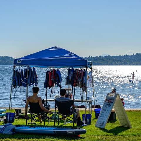 Color photo.  A blue-roofed pop-up shelter, next to a lake.  Two people are in its shade, sitting in front of a table, their backs to the camera, as they face the water.  Life vests hang from the shelter above them, and a stand up paddle board rests on the grass behind them.  Next to the shelter is a sign advertising rental rates for paddle boards.