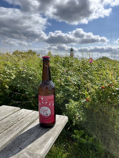 Bottle of Cykøl by Årø Bryg on the island of Årø, standing on a plate of wood. Behind it there“s greenery with an unfocused lighthouse and blue skies in the background.