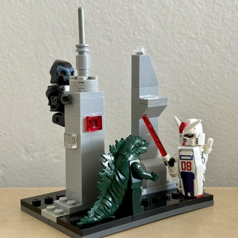 Three Lego Minifigures, Godzilla, Kong, and a Gundam, are in a small Lego city scene. Kong hangs from a skyscraper as Godzilla faces off against the Gundam.