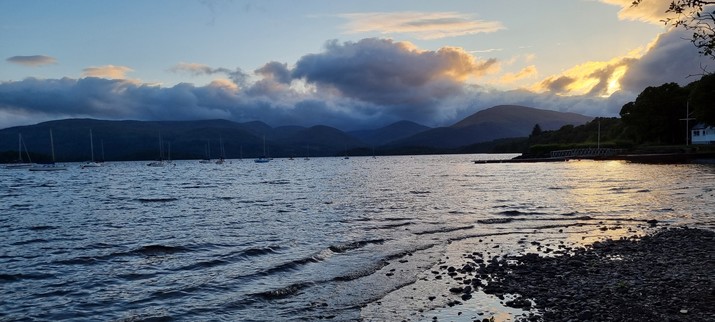 A photo of a view across Loch Lomond, taken from the east side. The sun is starting to set, but is mostly hidden behind clouds.