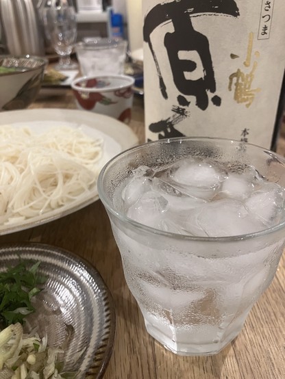 Glass of ice water, a bottle with Japanese text, a plate of noodles, and a small dish with garnishes on a table.