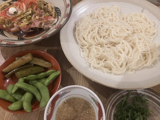 A spread of Japanese dishes including a bowl of noodles, a dish of vegetables (including edamame and pickles), a plate of cooked fish with sauce and garnish, a small bowl of dipping sauce, and a small portion of green herbs.