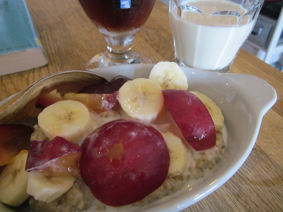 Close up on a dish of porridge with topping of sliced banana and plum.  A paper back book, a glass of iced coffee, and a glass of soy milk in the background