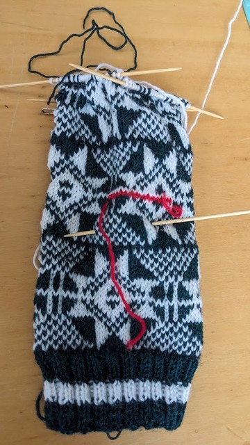 ４本の棒針を使って編んでいる途中の筒状の編み物の画像。深緑と白の糸が交互に使われていて幾何学模様が浮かび上がっています。途中に赤い毛糸が編まれています。
