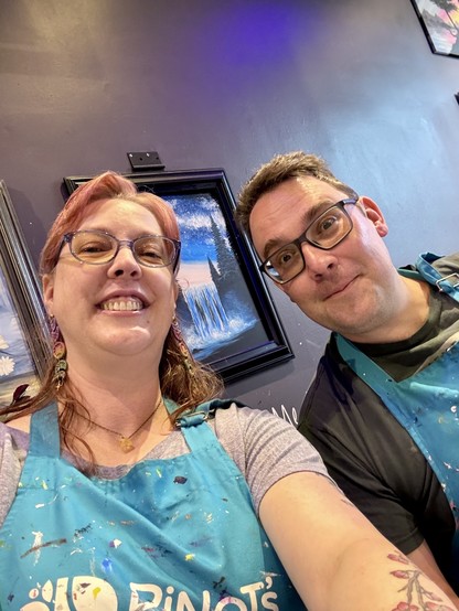 Two people wearing glasses and paint-splattered aprons, smiling at the camera. Artworks are visible in the background.
