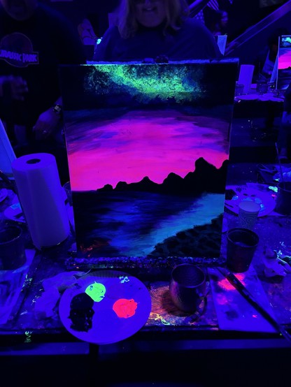 A neon painting of a landscape with mountains and water, illuminated under blacklight, displayed on an easel. Art supplies including paint, brushes, and cups are visible on a table around the painting. Two people are in the background.