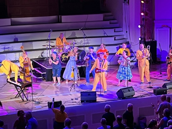 The Jive Aces and guests on stage, six men in matching zoot suits and an accordion-playing lady in a patterned dress. The keyboard player is partway through a handstand / mule kick on his instrument.