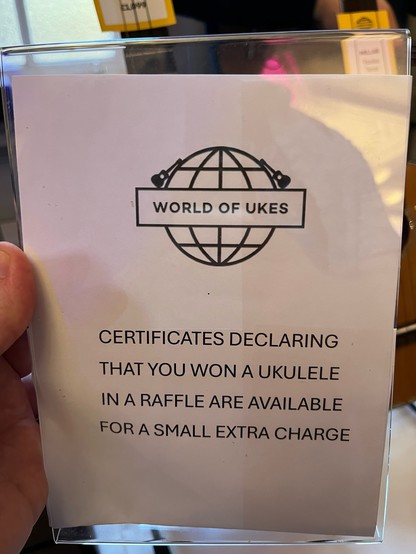 White sign with black text and globe logo. Text: WORLD OF UKES -
CERTIFICATES DECLARING THAT YOU WON A UKULELE IN A RAFFLE ARE AVAILABLE FOR A SMALL EXTRA CHARGE